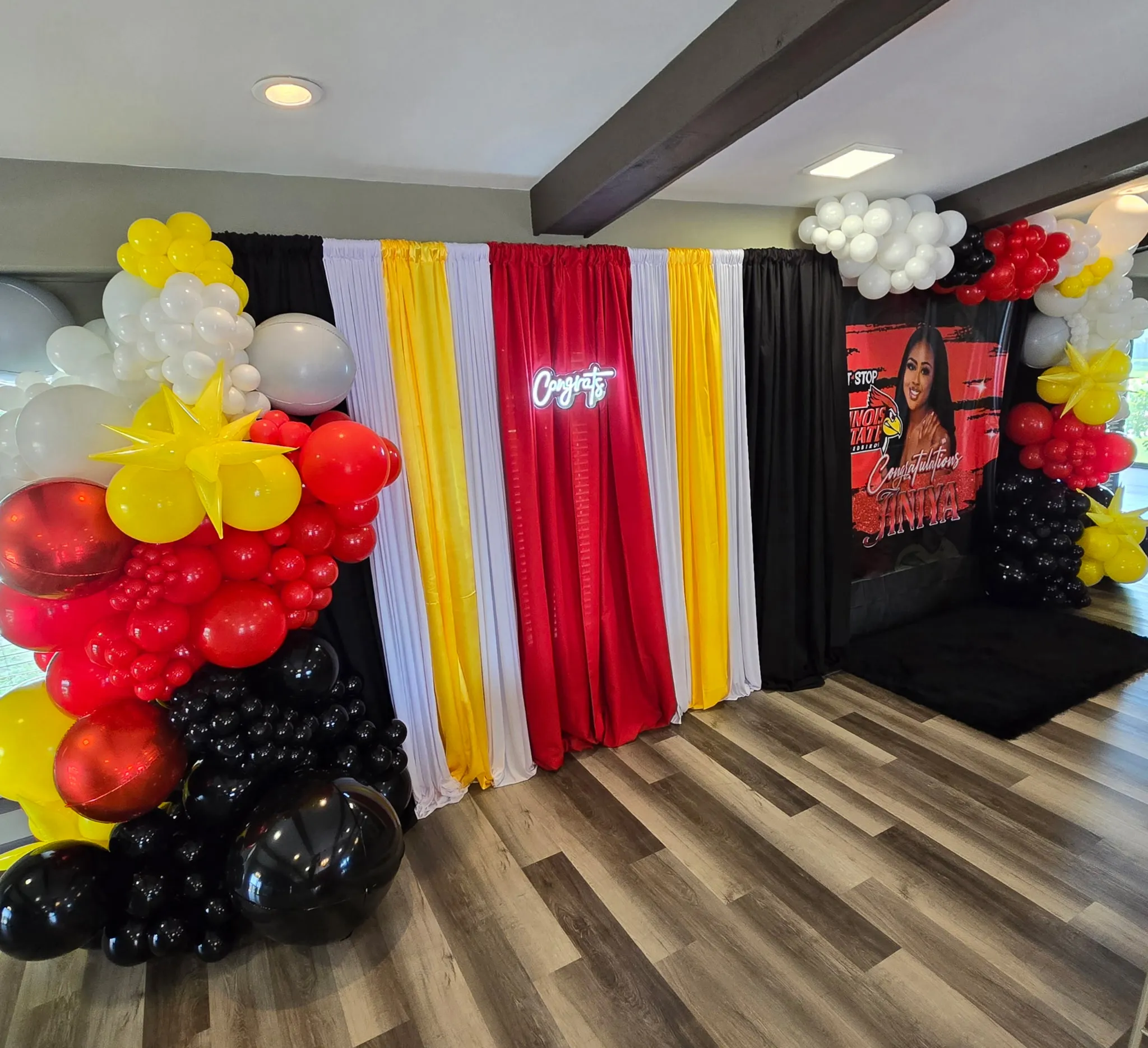 Graduation photo backdrop with draped fabric, balloon garlands, neon Congrats sign, and a custom graduation banner.